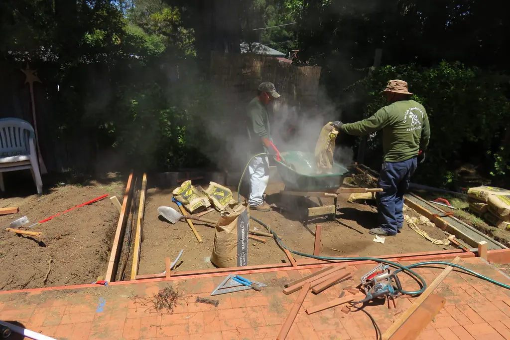 Two workers mixing bagged concrete beside the forms of a backyard pad