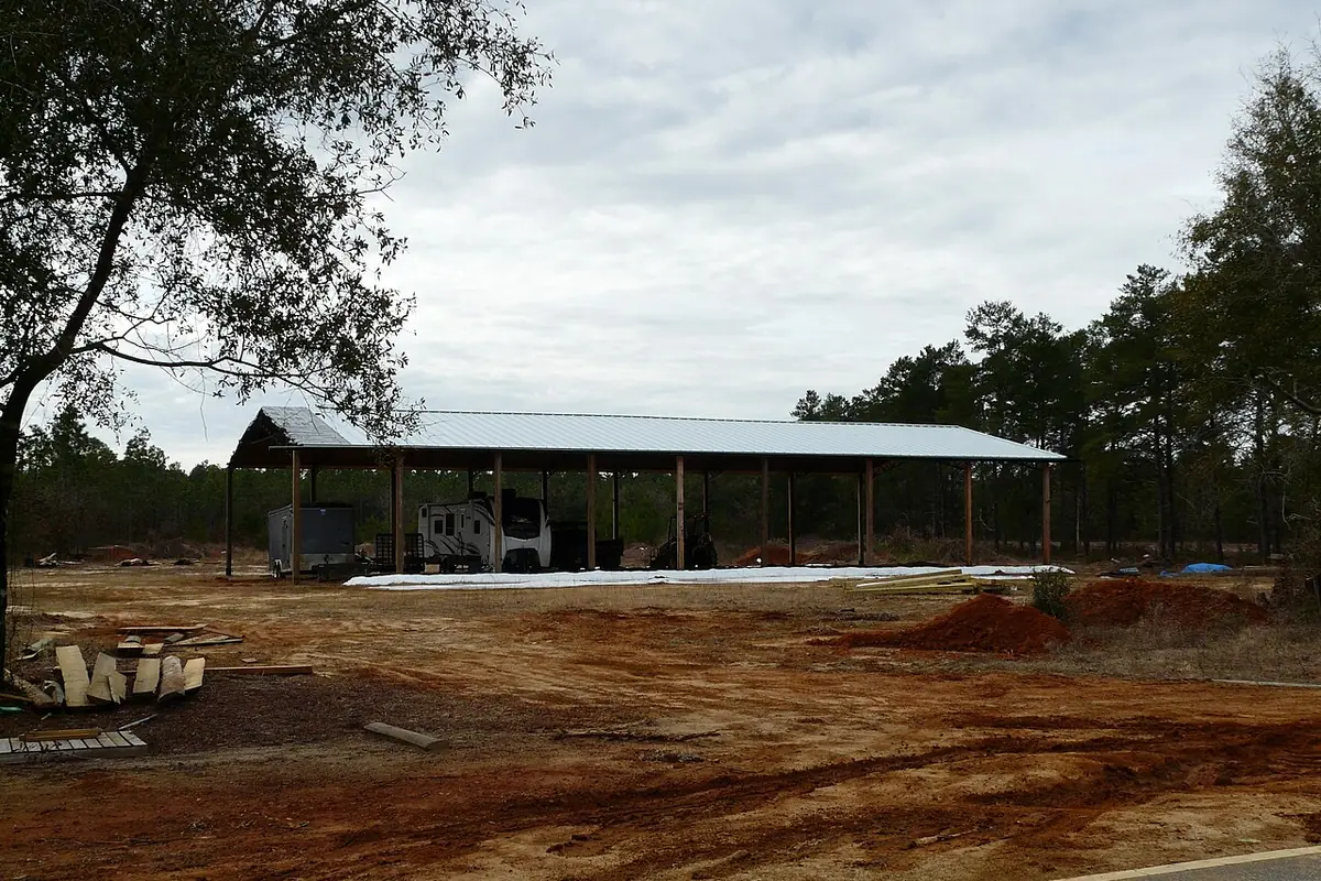Open pole barn shelter with an RV and equipment under the roof on a cleared lot