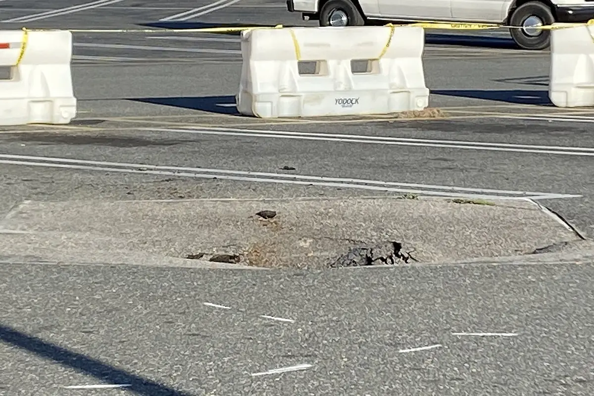 Cracked, sunken patch where a sinkhole reopened in a parking lot, fenced off with barriers
